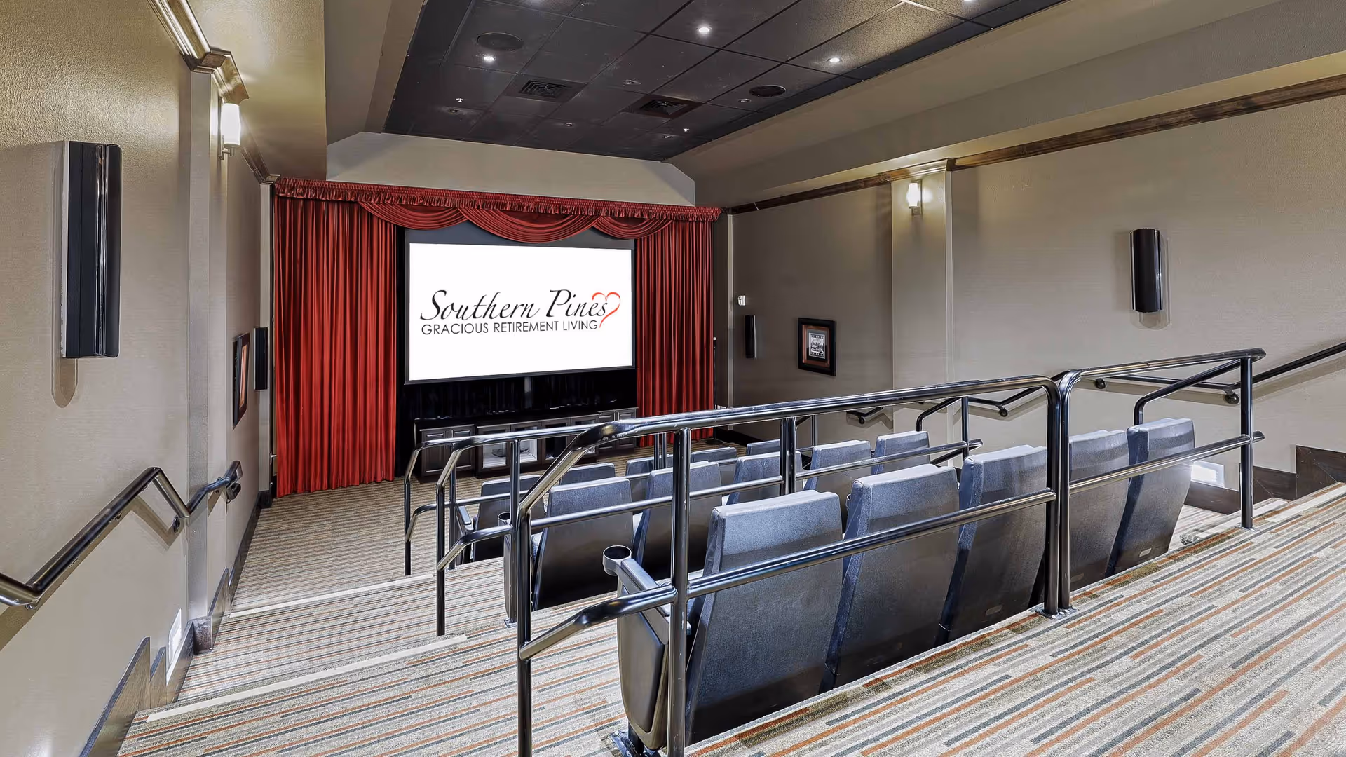 Interior view of a small theater room with tiered seating and handrails. The seats are upholstered in gray fabric and face a large screen framed by red curtains. The screen displays the logo for Southern Pines Gracious Retirement Living. The room has beige walls, wall-mounted speakers, and soft lighting fixtures.