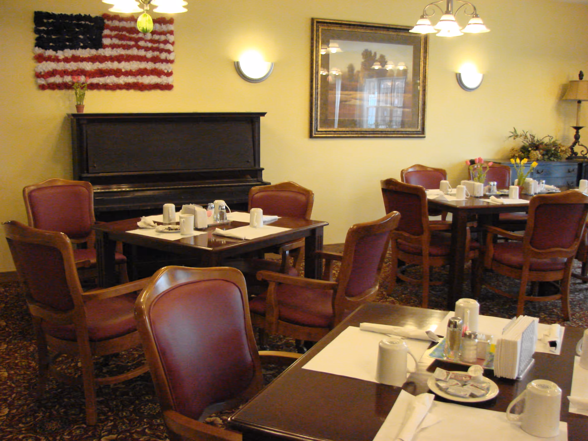 Dining room with several wooden tables and chairs with red cushions. Tables are set with white placemats, cups, napkins, and condiments. A black upright piano is against the wall with a decorative American flag made of flowers above it. A framed picture and wall-mounted lights are also visible on the yellow walls.