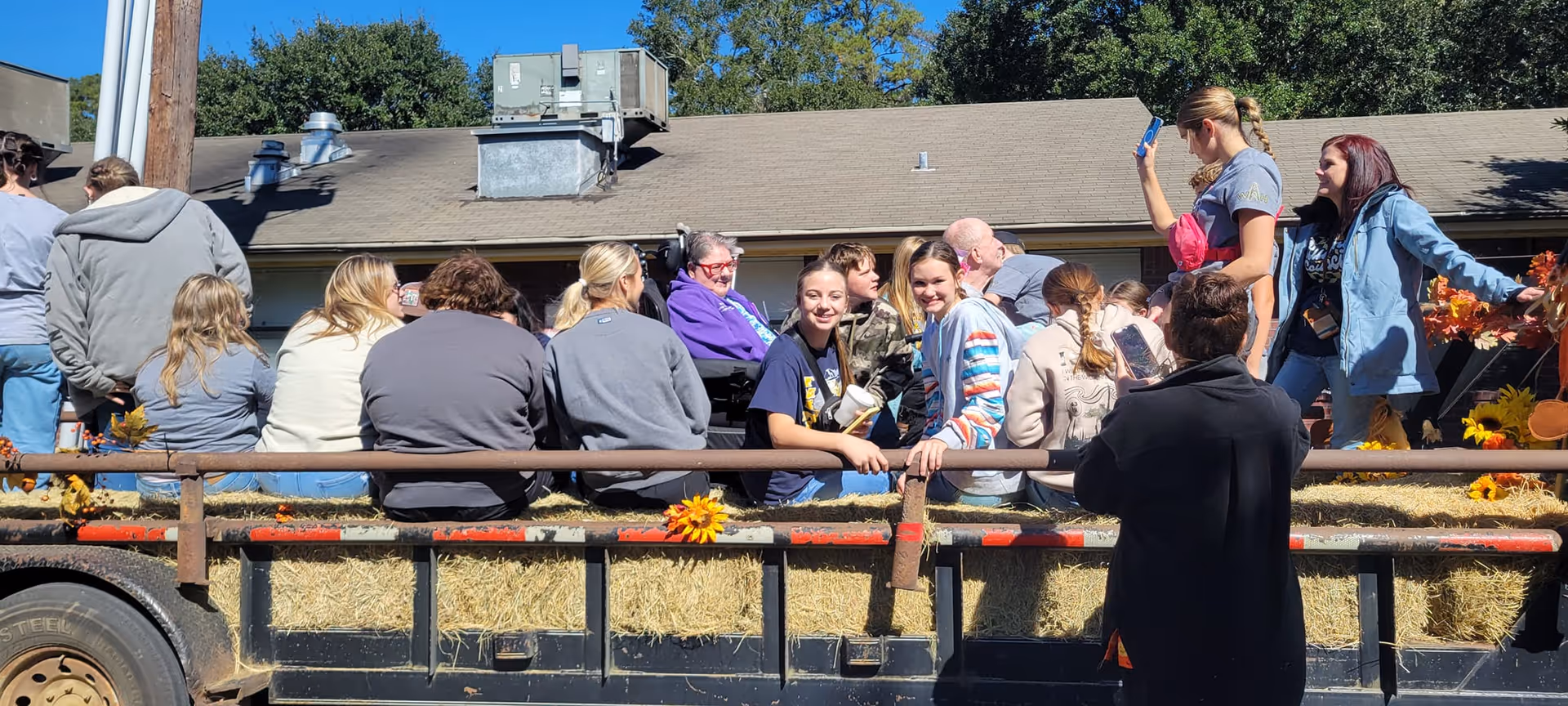 A group of people sitting on hay bales in the back of a flatbed trailer decorated with autumn-themed decorations, outdoors on a sunny day with a building and trees in the background.