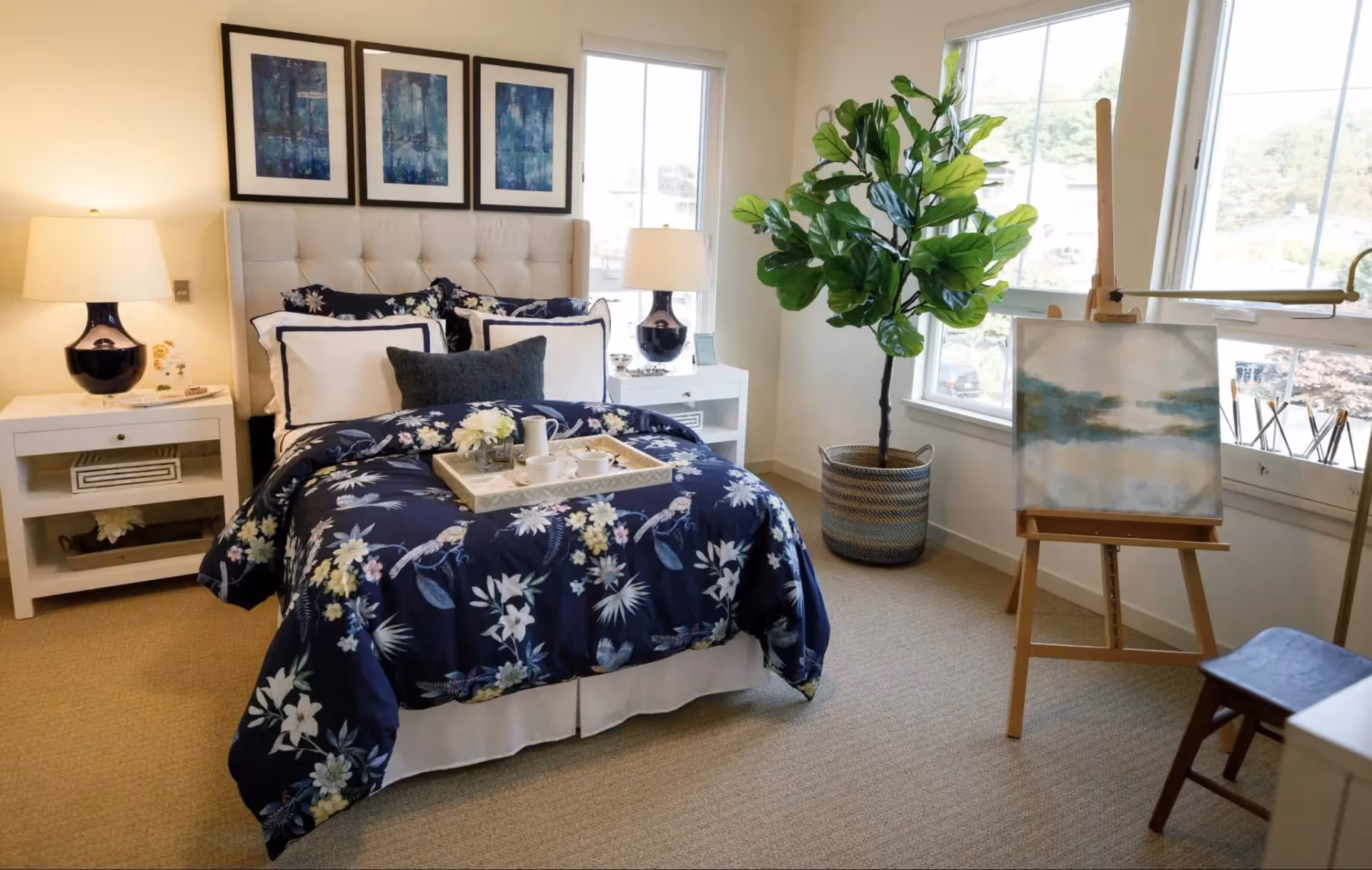 A cozy bedroom with a bed dressed in a dark blue floral comforter and multiple pillows. There are two white nightstands on either side of the bed, each with a lamp. Above the bed are three framed abstract art pieces. A large potted plant is placed near two windows letting in natural light. An easel with a painting and a small wooden stool are positioned near the windows.