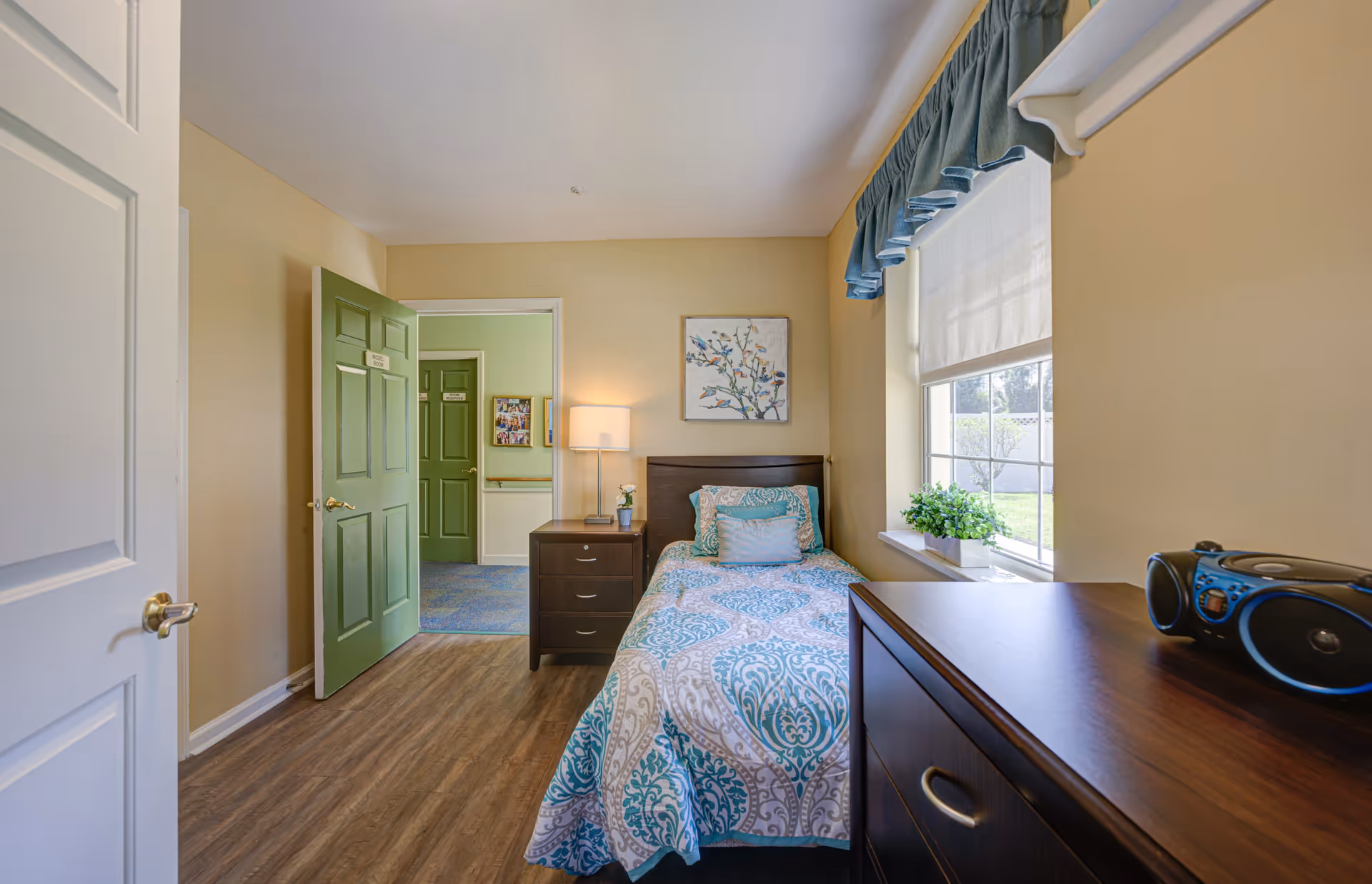 A small bedroom with a single bed covered in a blue and white patterned bedspread. Next to the bed is a dark wooden nightstand with a lamp and a small plant. A window with a blue valance and white shade lets in natural light. Across from the bed is a dark wooden dresser with a blue and black radio on top. The room has light beige walls and wood flooring. Two green doors are visible, one labeled 'Model Room'.