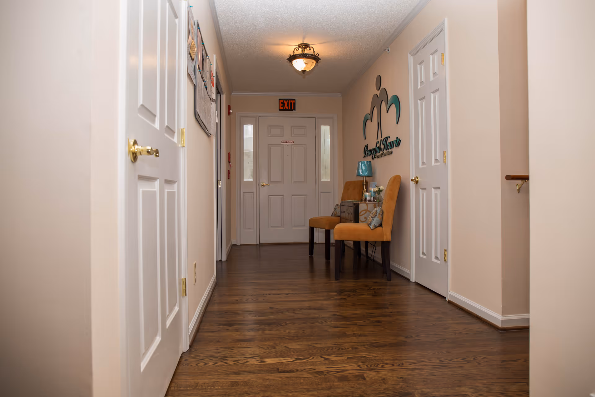 A hallway with wooden flooring and beige walls featuring several white doors on both sides. At the end of the hallway is a door with an exit sign above it. On the right side, there are two orange chairs with a small table between them holding a lamp and decorative items. Above the chairs is a wall decoration with the text 'Peaceful Hearts Personal Care Home'.