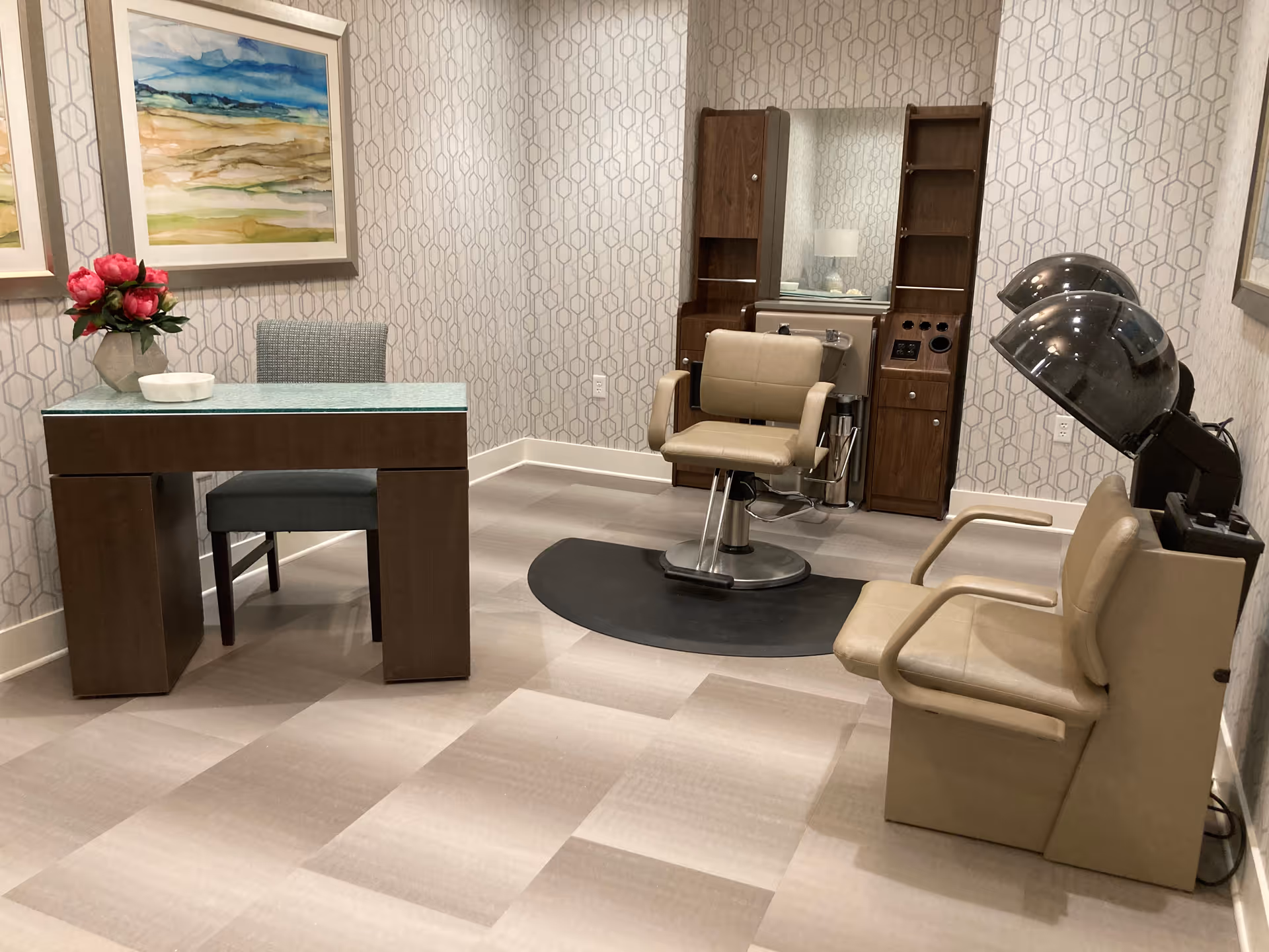 Interior view of a salon room with a styling chair in front of a mirror and wooden cabinetry, a hair dryer chair with two hooded dryers, a desk with a chair, a vase with pink flowers, and framed artwork on the wall.