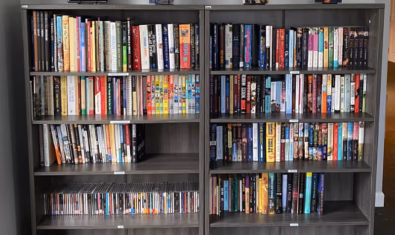 Two dark wooden bookshelves filled with a variety of books and a collection of CDs on the bottom shelf, placed against a wall in an indoor setting.