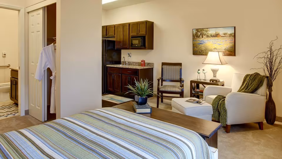 A cozy senior living studio apartment featuring a bed with a striped bedspread in the foreground, a small kitchenette with dark wood cabinets and a black refrigerator, a sitting area with a white armchair and ottoman draped with a green throw, a wooden chair, a side table with a lamp and decorative birds, a framed landscape painting on the wall, and a bathroom visible through an open door with a white robe hanging on the closet door.