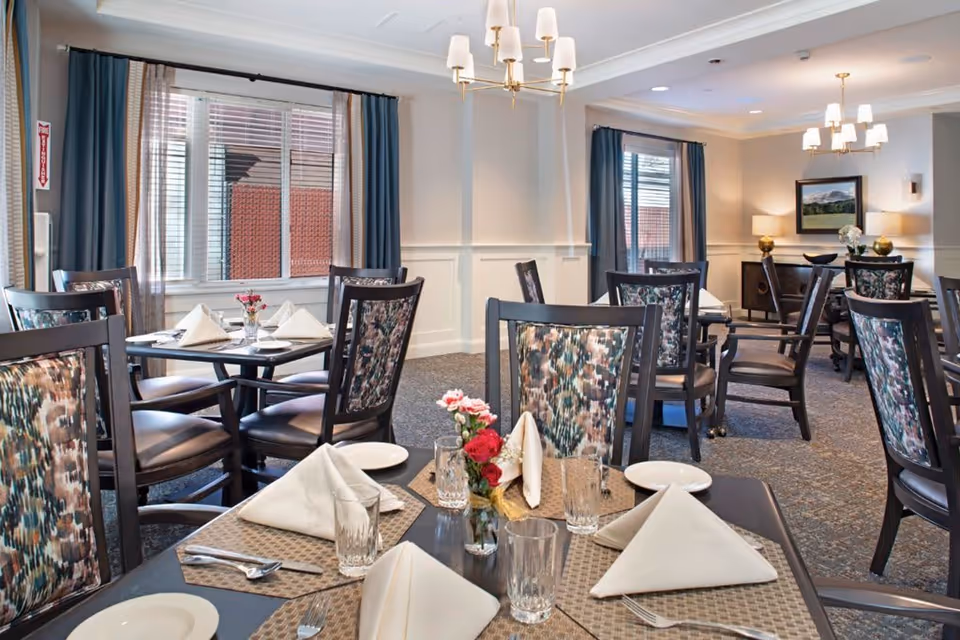 Bright dining room with multiple set tables, upholstered patterned chairs, chandeliers, and windows.