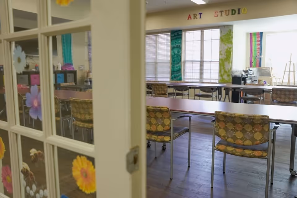 View through a glass-paneled door decorated with flower stickers into an art studio room with tables and patterned chairs. The room has large windows with colorful fabric panels and the words 'ART STUDIO' displayed above the windows.