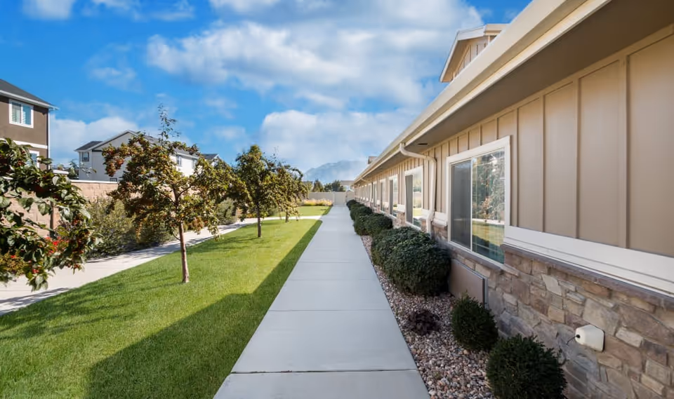 Sidewalk runs between a single-story building with windows and landscaped grass, shrubs, and trees under a blue sky.