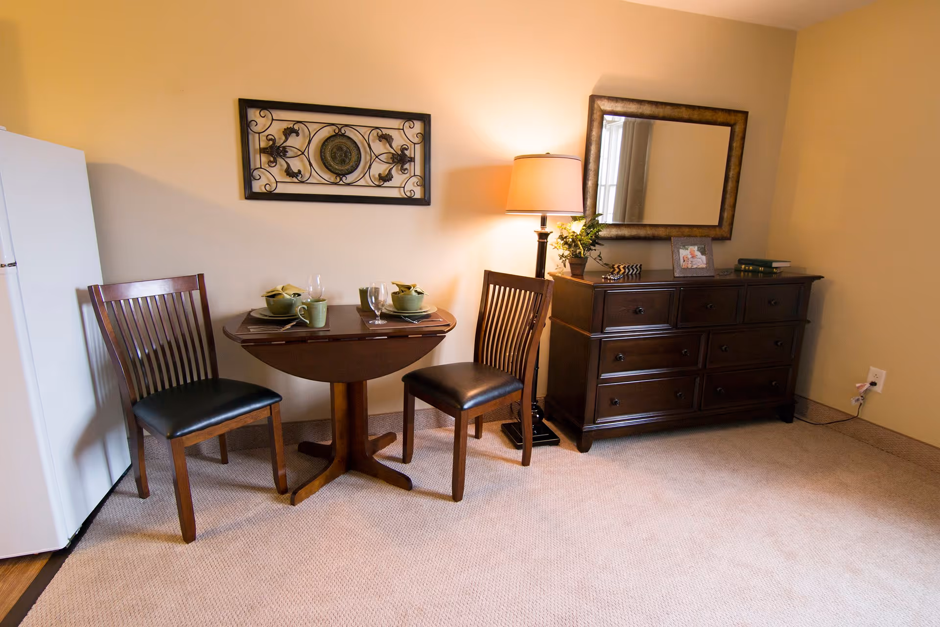 A small dining nook with a round wooden table set for two, two wooden chairs, a floor lamp, and a dark wood dresser topped with a mirror.