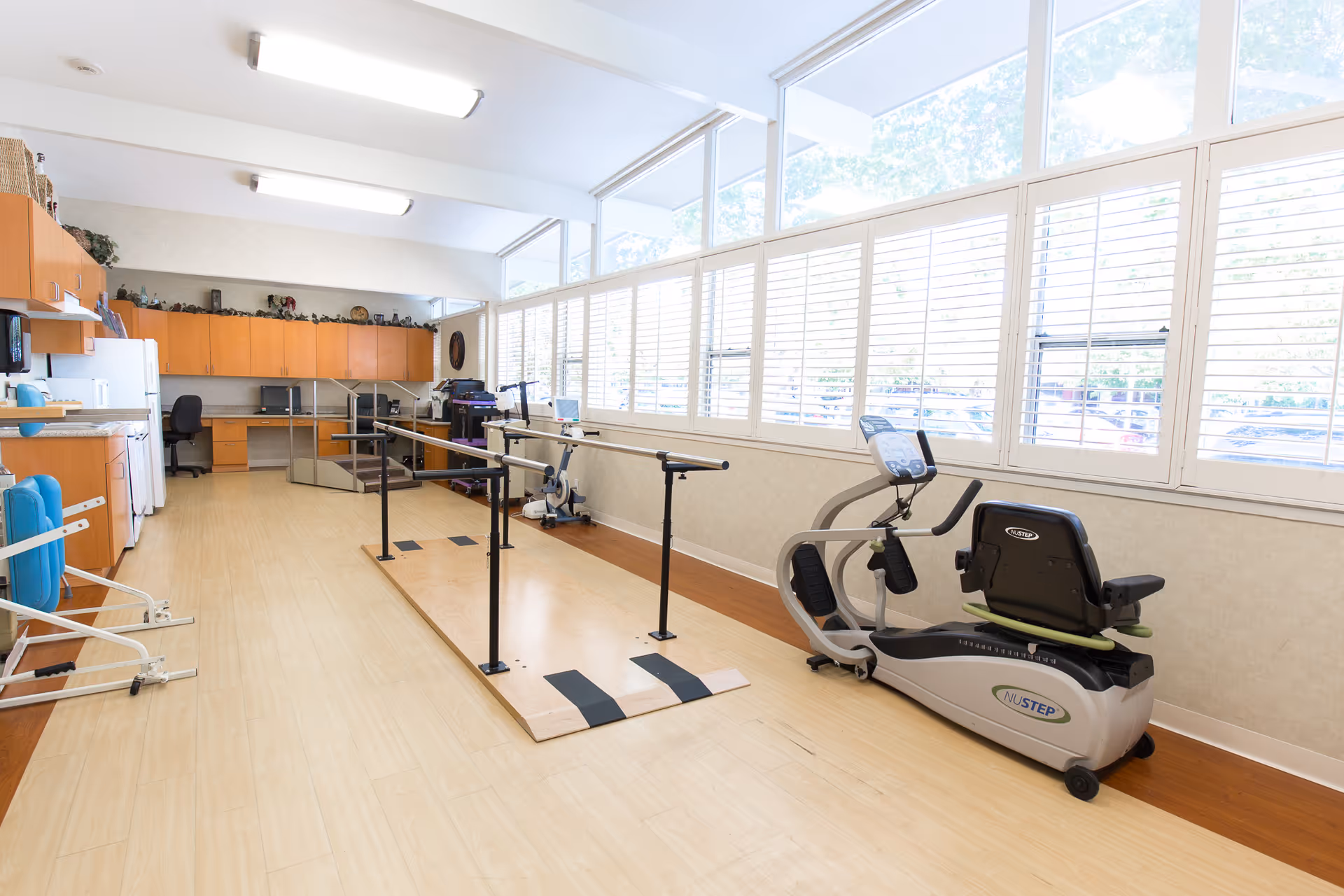 A bright rehabilitation room with large windows covered by white blinds. The room features parallel bars for walking exercises, a NuStep recumbent cross trainer, and a desk area with office chairs and cabinets along the back wall. The floor is light wood, and the space is well-lit with ceiling lights.