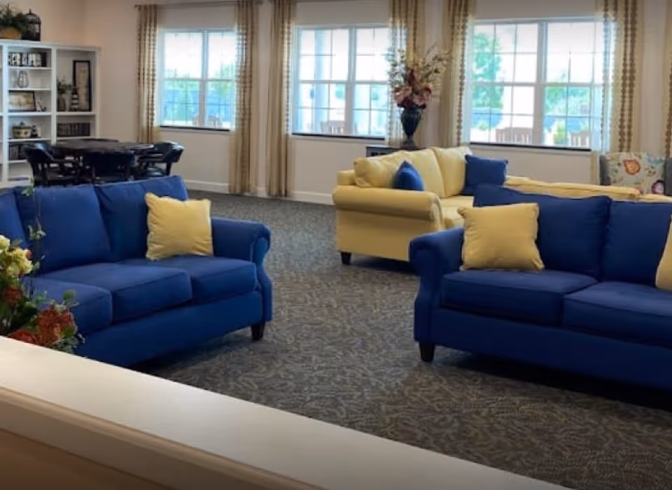 A bright living room area with two blue sofas and one yellow sofa, each adorned with yellow and blue throw pillows. Large windows with beige curtains allow natural light to fill the room. In the background, there is a dining table with chairs and a white bookshelf filled with decorative items and books. A vase with flowers is placed on a table near the windows.