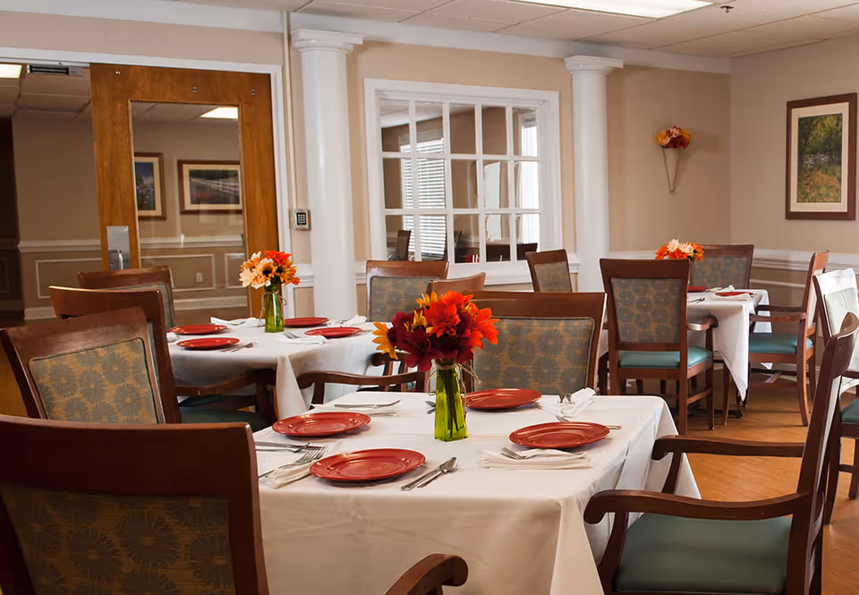 A dining room with several tables covered in white tablecloths, each set with red plates, silverware, and napkins. Each table has a small green vase with colorful flowers. The room has wooden chairs with patterned cushions, beige walls, framed artwork, and a large window with white trim.