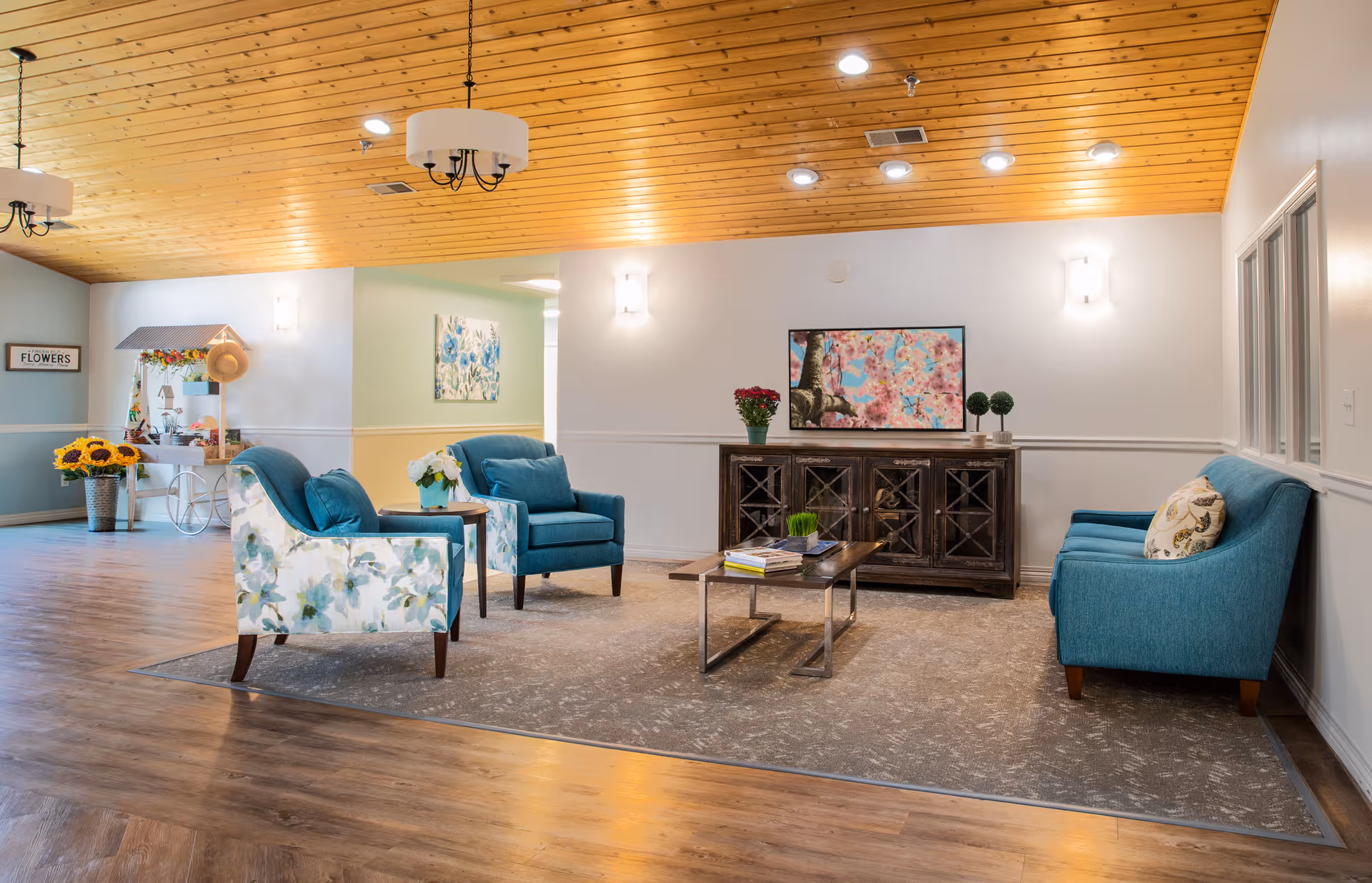 Light-filled common lounge with teal and floral armchairs arranged around a coffee table under a wooden ceiling.