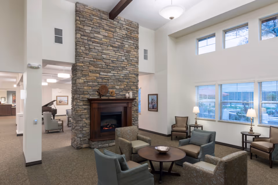 A cozy living room area with a tall stone fireplace as the focal point. The room features multiple armchairs arranged around a round wooden coffee table. There are large windows letting in natural light, with two table lamps on side tables near the windows. The ceiling is high with exposed wooden beams and a hanging light fixture. In the background, there is a glimpse of another room with a piano and additional seating.