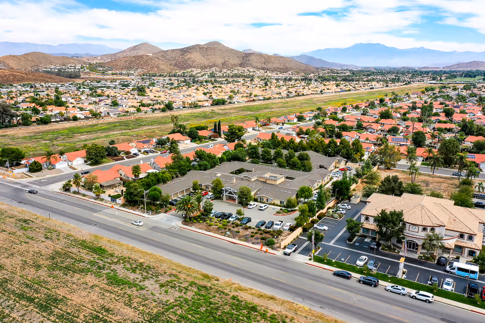 Aerial view of the Sunny Rose Senior Living complex and surrounding suburban neighborhood with parking lots, a main road, and distant hills.