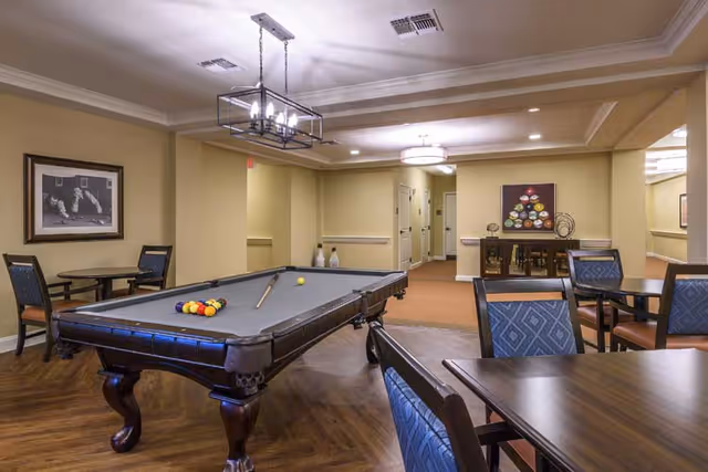 A senior living common room with a pool table, multiple seating tables and chairs, and overhead lighting.