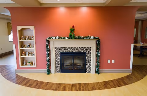 A cozy indoor space featuring a fireplace with a tiled surround and a white mantel decorated with garland and a small Christmas tree. The wall behind the fireplace is painted a warm red color. To the left of the fireplace is a built-in shelf displaying various small decorative figurines. The floor has a light wood finish with a darker wood curved inlay pattern. The room appears well-lit with ceiling lights.