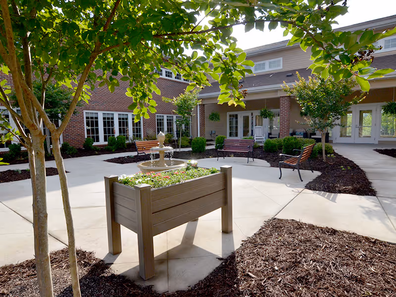 Sunlit courtyard with a raised planter, central fountain, benches, and the building's brick facade framed by trees.