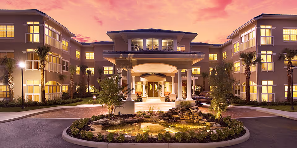 Exterior view of a large senior living facility building at sunset with warm lights glowing from the windows. The entrance features a covered driveway with white columns and a landscaped roundabout with a small pond and rocks in front.