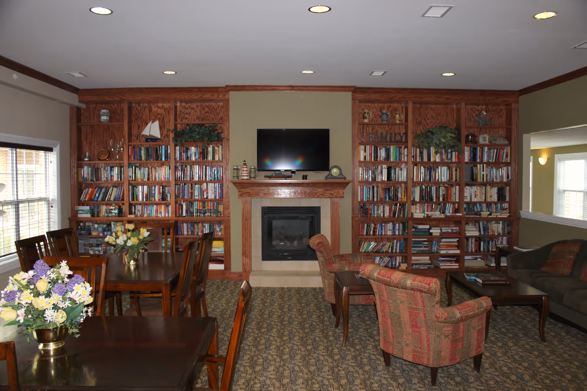 A cozy living room area with built-in wooden bookshelves filled with books and decorative items. A flat-screen TV is mounted above a fireplace in the center. There are two patterned armchairs and a coffee table in front of a sofa on the right side. On the left, there are wooden dining tables with flower arrangements. The room has carpeted flooring and recessed ceiling lights.
