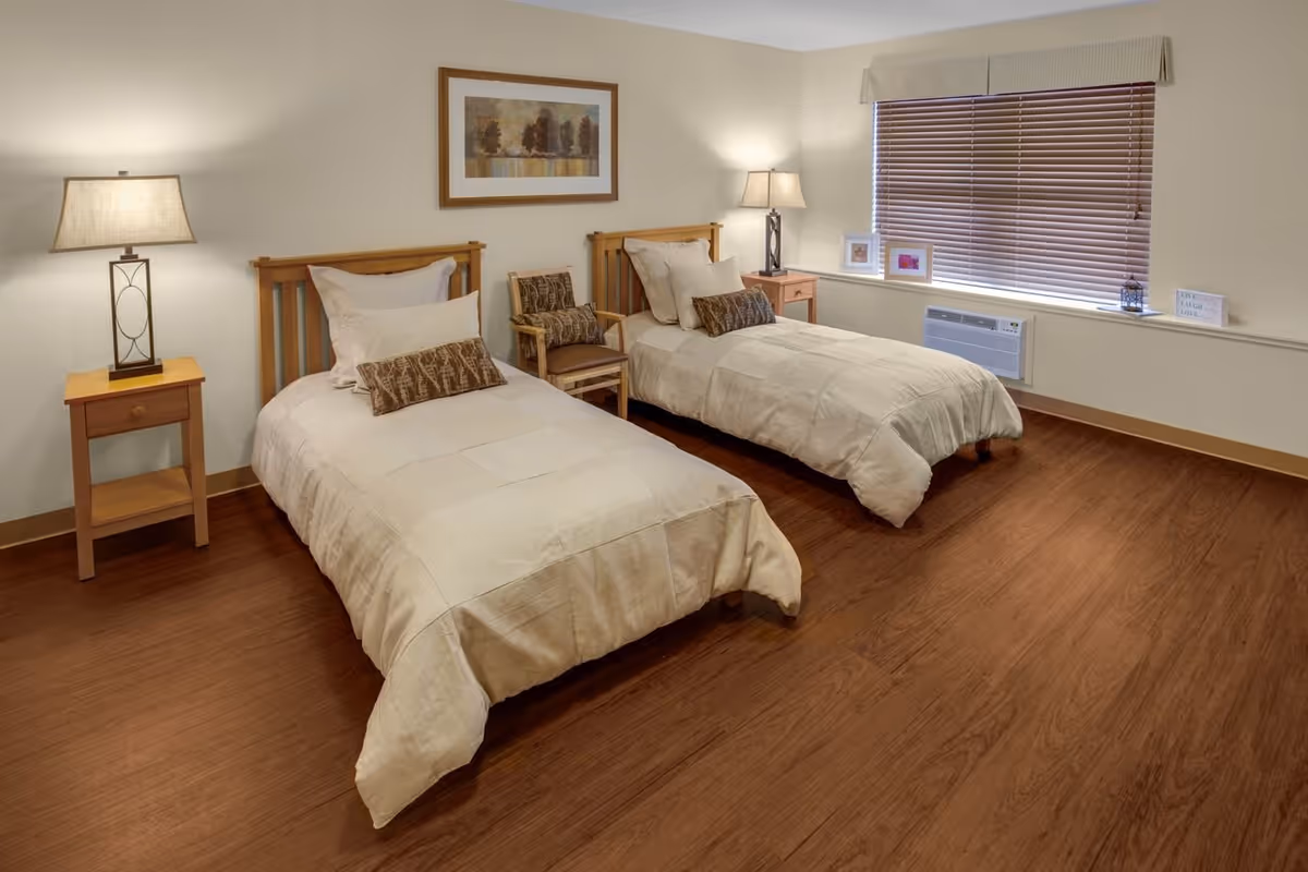 A bedroom with two twin beds, wooden headboards, nightstands with lamps, hardwood floors, and a window with blinds.