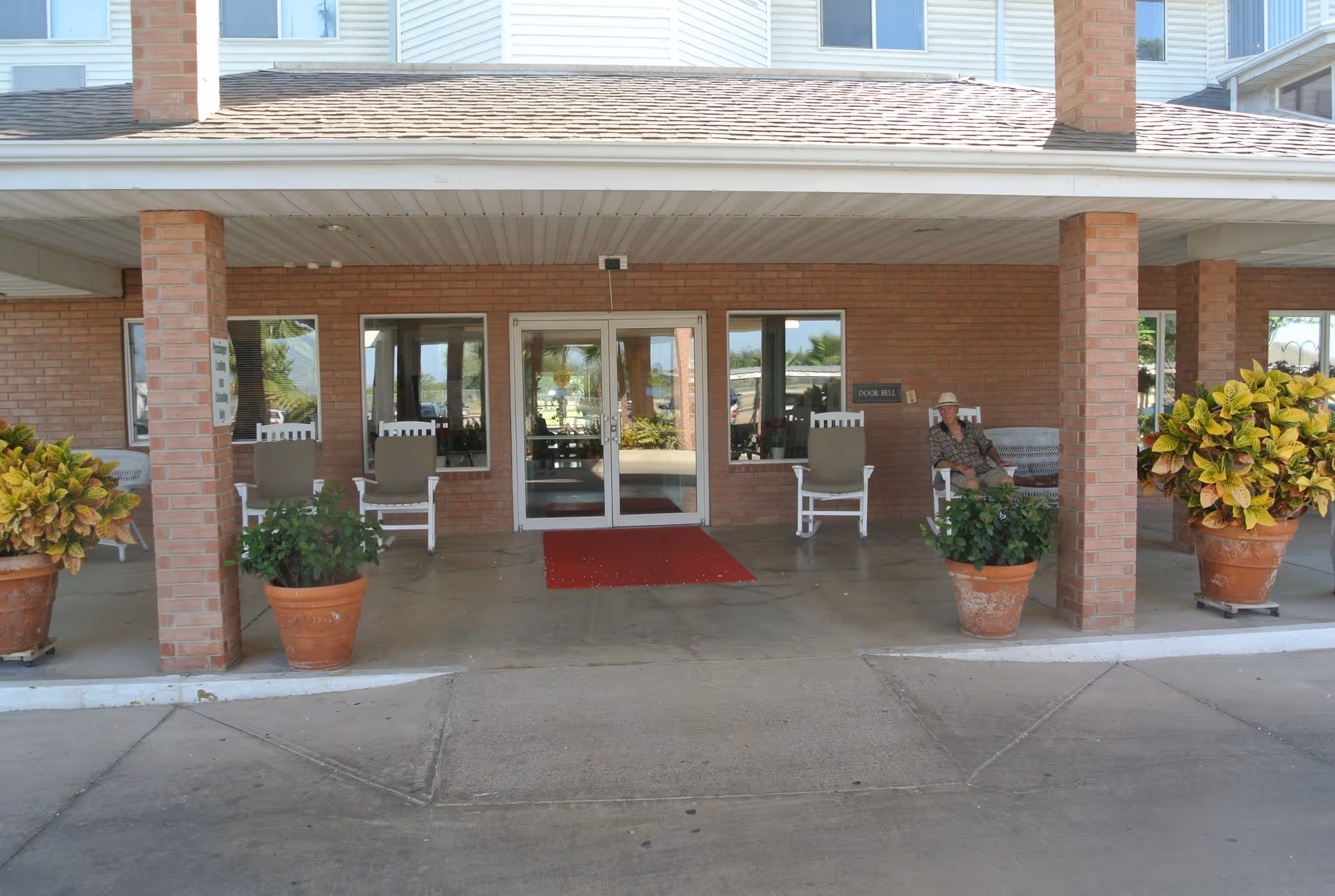 Entrance area of a retirement community building with a covered porch supported by brick columns. There are several potted plants and rocking chairs on the porch. A person wearing a hat is sitting on one of the chairs near the right side. The entrance has double glass doors with windows on either side and a red doormat in front.