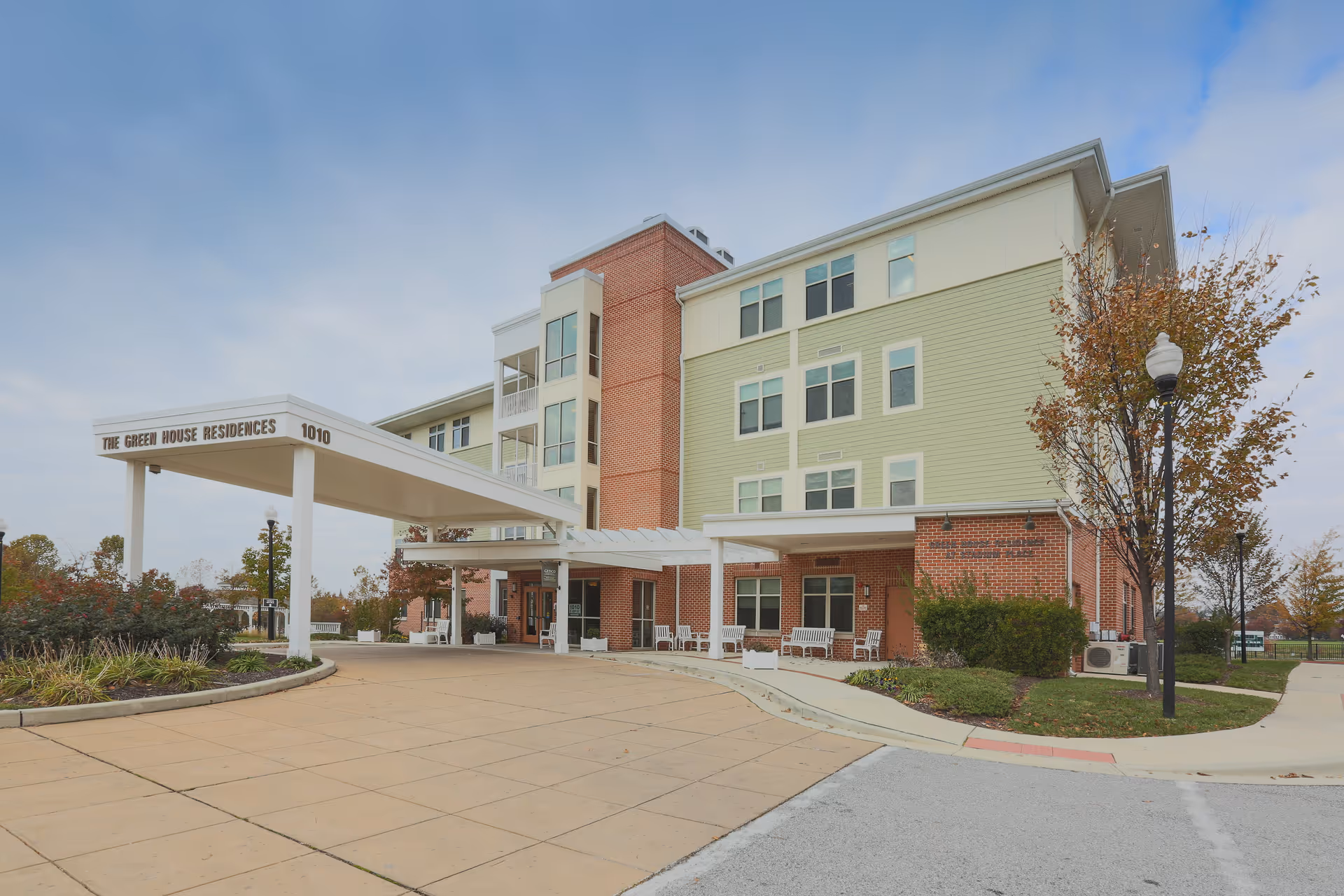 Front exterior of a multi-story brick and pale-green senior residence with a covered entrance canopy reading 'The Green House Residences 1010' and benches by the driveway.