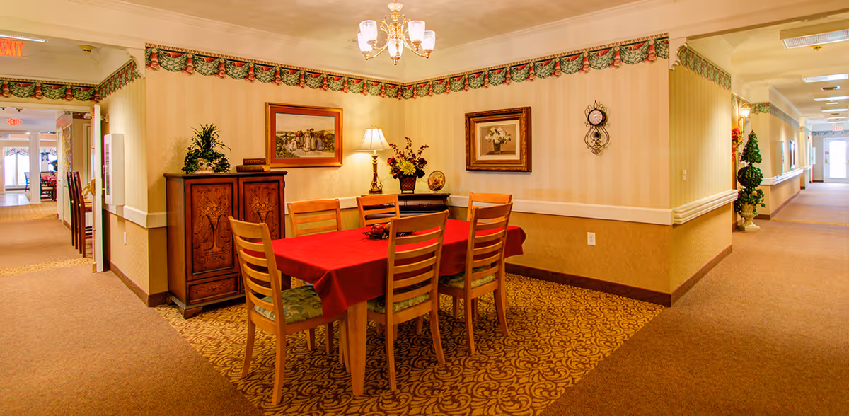 A cozy dining area in a senior living facility with a rectangular wooden table covered by a red tablecloth and six wooden chairs with patterned cushions. The walls have beige striped wallpaper with a decorative floral border near the ceiling. There are framed paintings, a wall clock, a table lamp, and flower arrangements on wooden cabinets against the walls. The area is carpeted with a patterned carpet and is adjacent to a hallway with beige carpeting and multiple exit signs.