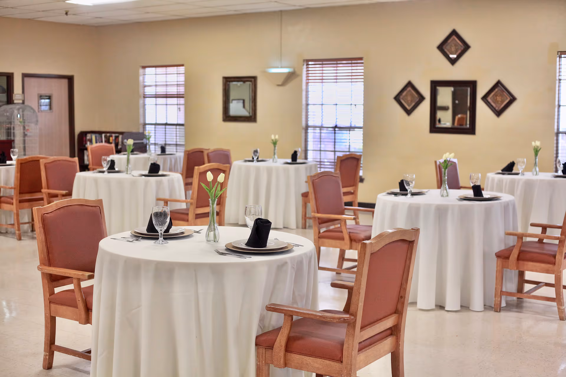 A dining room in a nursing and rehab center with round tables covered in white tablecloths, each set with plates, glasses, black napkins, and small vases holding white tulips. The room has wooden chairs with padded seats and backs, beige walls with decorative mirrors, and windows with blinds letting in natural light.