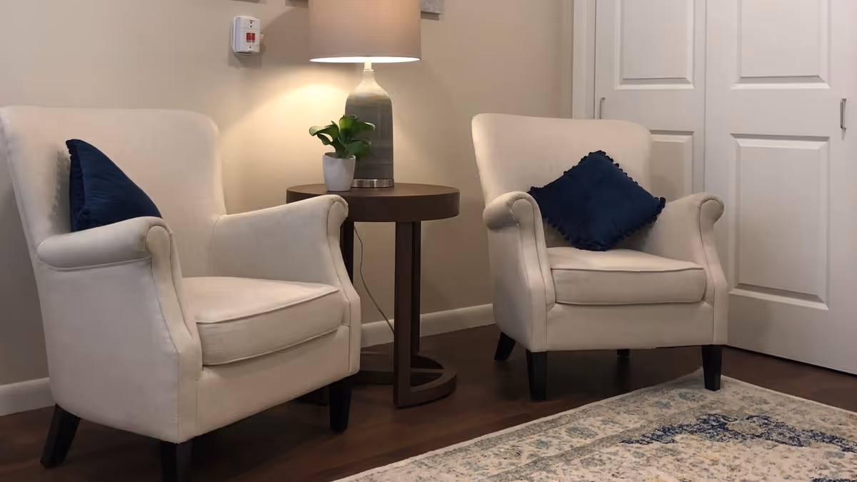Two beige armchairs with blue throw pillows flanking a round side table topped with a lamp and small plant in a cozy interior seating area.