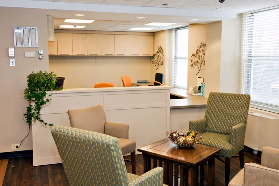 A cozy reception or waiting area with a white reception desk in the background, two orange chairs behind the desk, and a small seating area in the foreground with four upholstered chairs around a wooden coffee table holding a decorative bowl. The room has large windows with blinds and light beige walls decorated with metal wall art and a green potted plant on the left side of the desk.