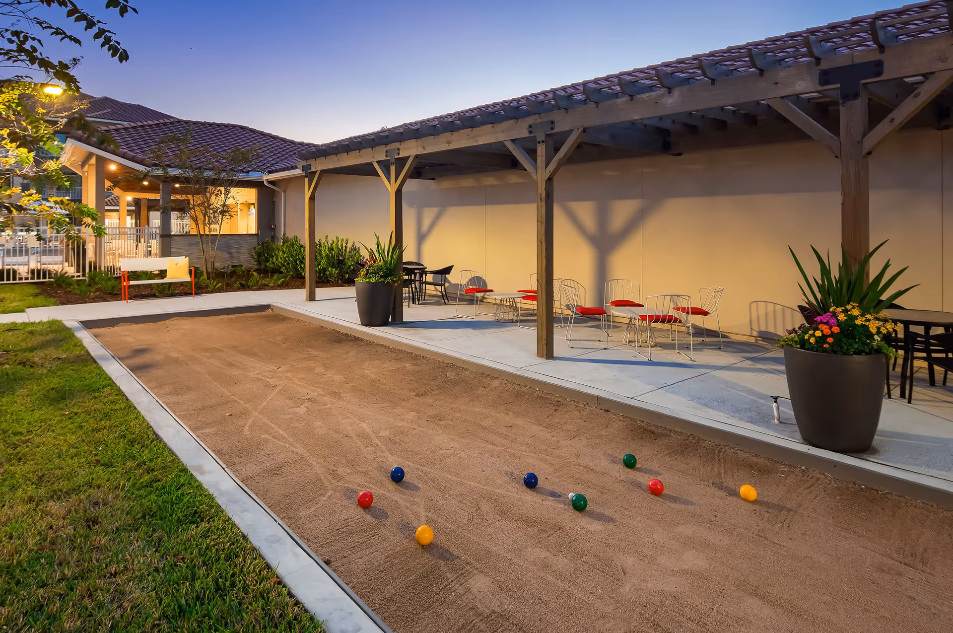 Outdoor bocce ball court with colorful bocce balls on the sand. There is a covered seating area with metal chairs and tables, some with red cushions. Large potted plants with flowers are placed near the seating area. The scene is set during dusk with a building featuring a tiled roof and warm lighting in the background.
