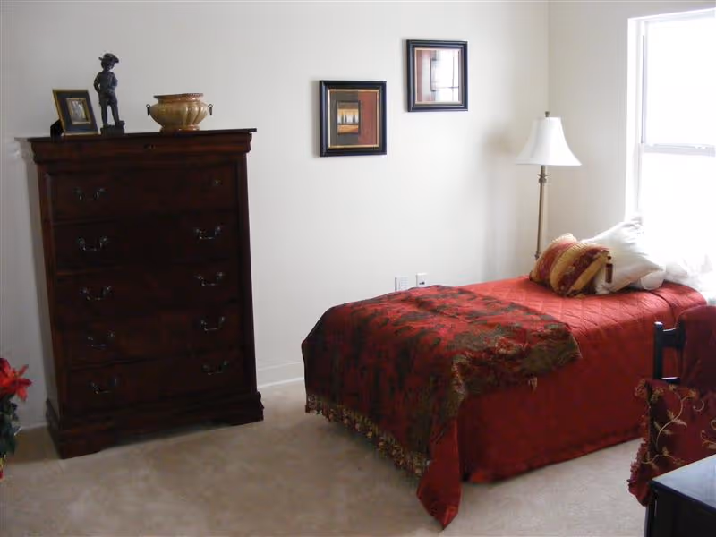 A bedroom with a single bed covered in a red and brown patterned bedspread, several pillows, a wooden chest of drawers with decorative items on top, two framed pictures on the wall, a floor lamp, and a window letting in natural light.
