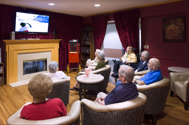 A group of elderly people seated in a cozy living room watching a television mounted above a fireplace. The room has wooden flooring, red curtains, a popcorn machine, and comfortable armchairs arranged in a semi-circle facing the TV.