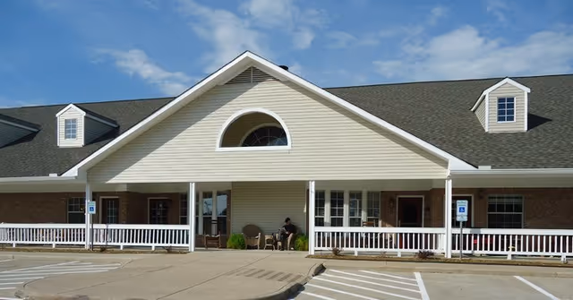 Front exterior view of a single-story building with a large covered porch, white railings, and two handicap parking spaces in front. The building has a beige siding and brick facade with a dark shingled roof and two dormer windows. A person is sitting on the porch.