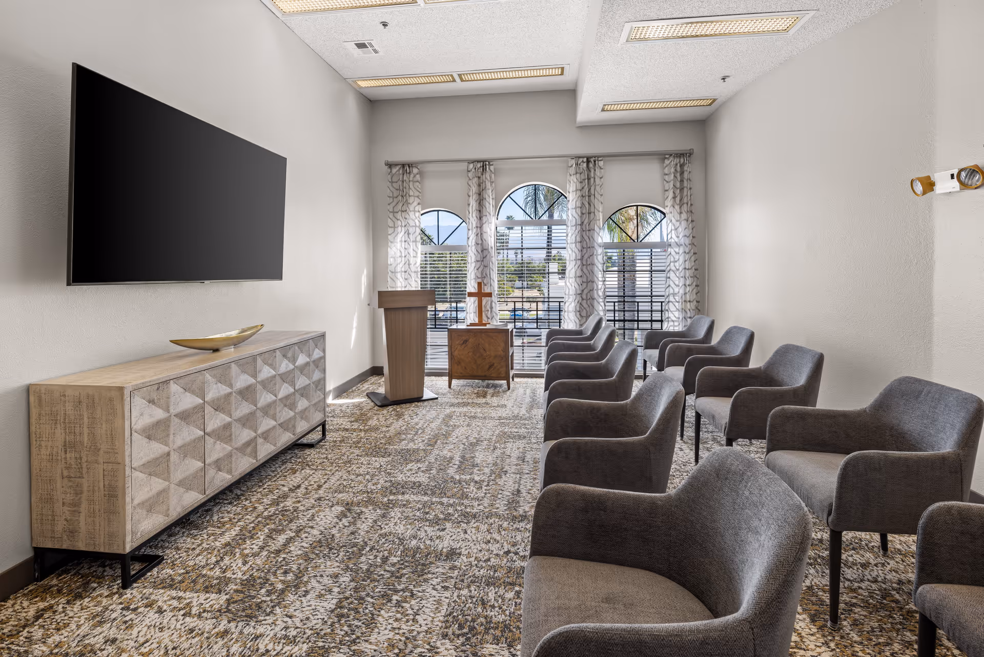 A small meeting or chapel room with rows of gray upholstered chairs facing a wooden podium and a small table with a cross. The room has three arched windows with patterned curtains, a large flat-screen TV mounted on the wall, and a textured wooden cabinet beneath the TV. The floor is carpeted with a patterned design.
