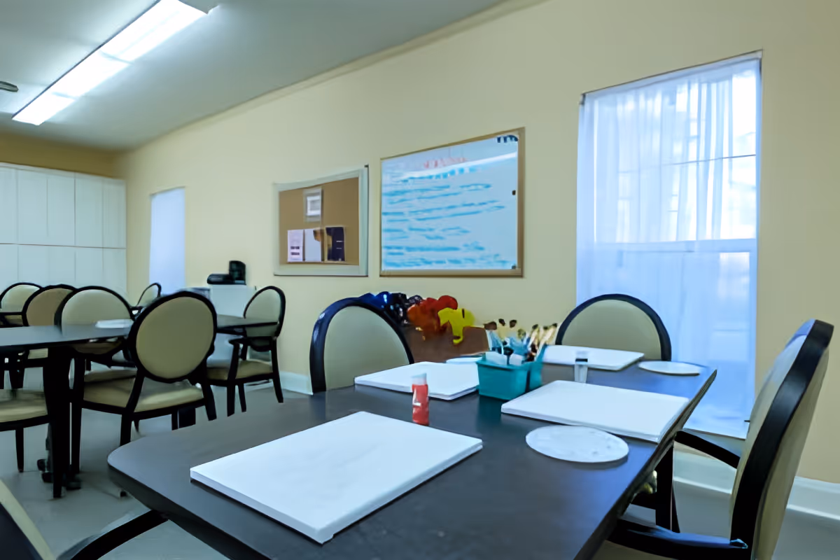 Bright activity room with several tables and round-backed chairs, place mats and art supplies on the foreground table.