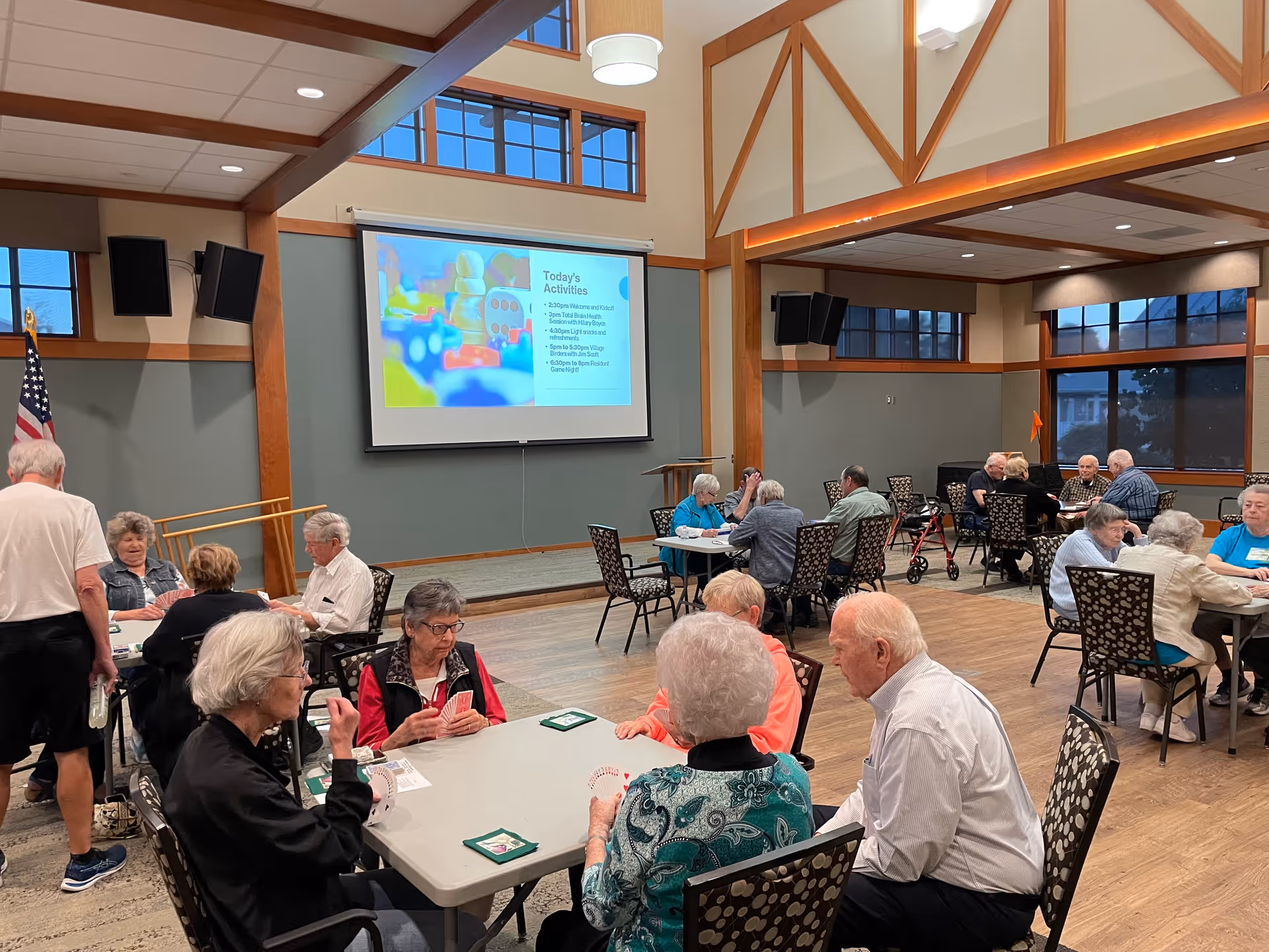 A group of elderly people sitting at tables in a large, well-lit community room playing card games. The room has high ceilings with wooden beams, large windows, and a projector screen displaying today's activities. An American flag is visible in the corner.