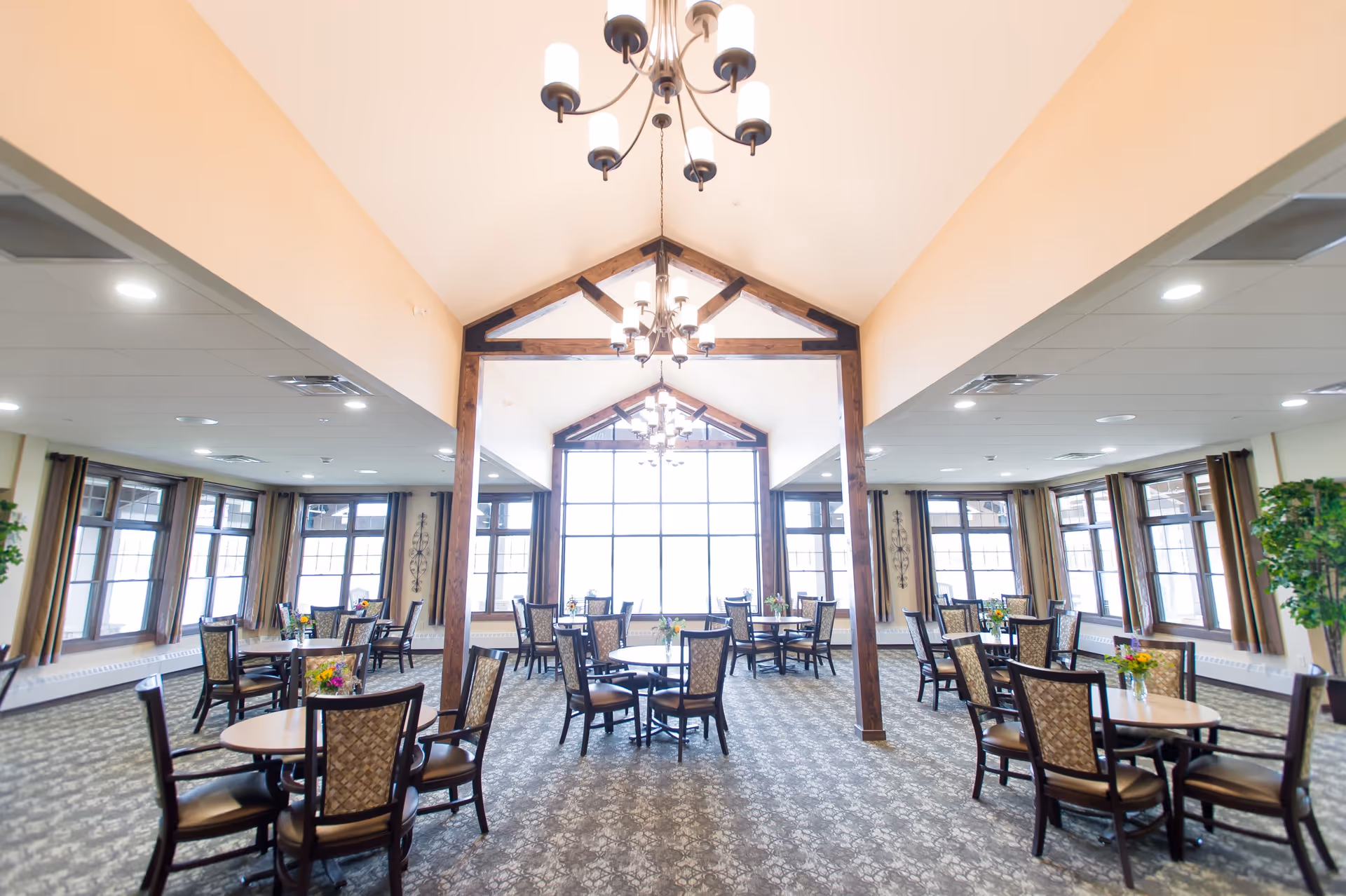 Spacious senior dining room with multiple round tables and chairs, large windows, chandeliers, and exposed wooden beams.