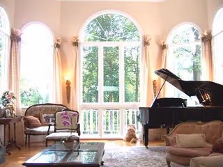 Sunlit living room with a grand piano, large arched windows, plush seating, and a glass-top coffee table.