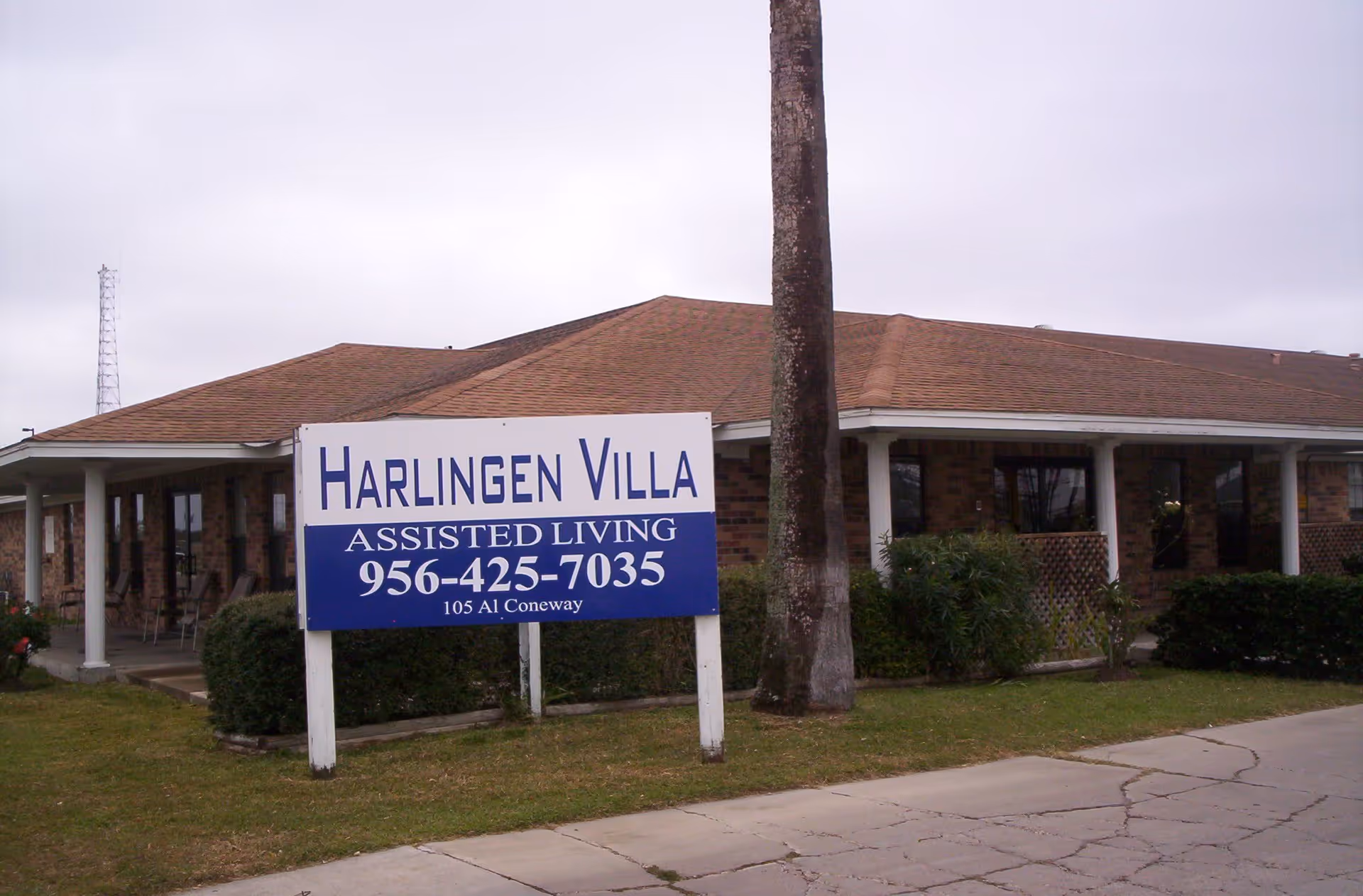 Exterior view of a single-story brick building with a brown shingled roof and a covered porch area. In front of the building is a large sign that reads 'Harlingen Villa Assisted Living 956-425-7035 105 Al Coneway'. There is a palm tree and some bushes near the sign and building.
