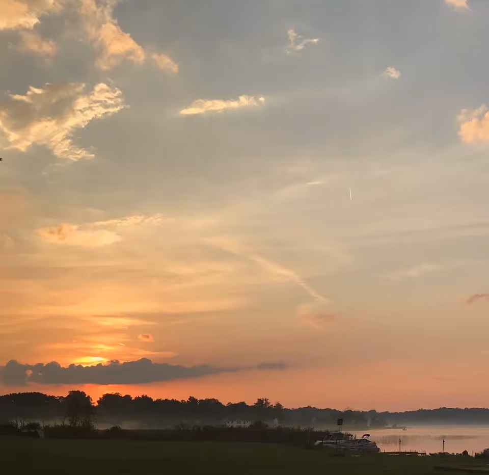 Sunset over a calm lake with a partly cloudy sky. Trees and a grassy area are visible in the foreground, with boats docked along the shoreline to the right.