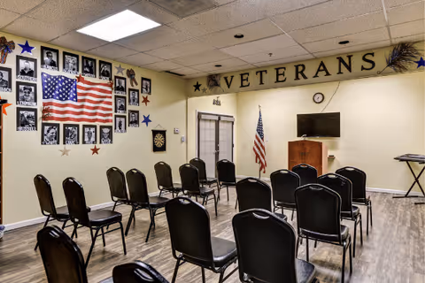 A room set up with rows of black chairs facing a podium and a wall-mounted TV. The wall behind the podium has the word 'VETERANS' displayed with stars on either side. An American flag is positioned next to the podium. Another wall features framed photos of veterans with an American flag and decorative stars.