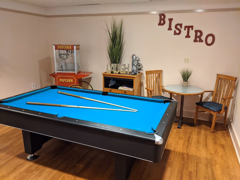 A recreational room with a blue felt pool table with two pool cues resting on it. In the background, there is a popcorn machine, a small wooden cabinet with decorative items including a plant and a 'LAUGH' sign, and a small round table with two wooden chairs. The word 'BISTRO' is displayed on the wall above the table.