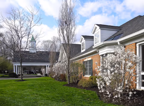 Exterior view of a senior living facility with a brick building, white flowering bushes, leafless trees, and a green lawn under a partly cloudy sky.