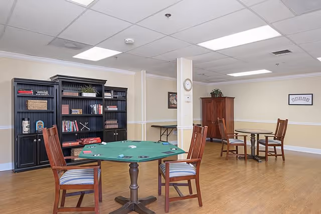 A spacious room with wooden flooring featuring two round tables with chairs around them. One table has a green felt surface with playing cards and poker chips, suggesting a game area. The room also contains black bookshelves filled with books and decorative items, a wooden cabinet, and a clock on a white pillar. The walls are painted beige with white trim, and the ceiling has recessed lighting.