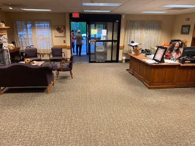 Reception area of a retirement community with a wooden front desk on the right where a woman is seated working on a computer. There are two people near the glass entrance doors at the back. On the left, there is a seating area with purple upholstered chairs and a couch, a small table with a sunflower decoration, and a fireplace. The room has beige walls and carpeted floor with windows covered by light curtains.