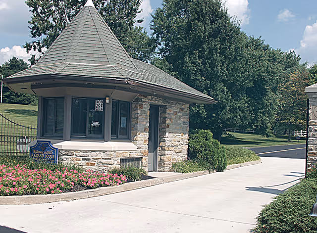 Stone guardhouse with a conical roof at the entrance of a driveway, surrounded by green trees and pink flowers, with a sign reading 'Masonic Homes & Grand Lodge'.