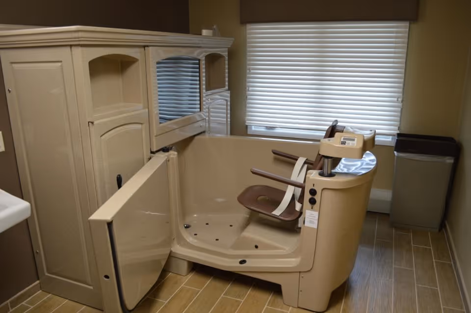 A specialized bathing chair and tub unit designed for assisted bathing in a senior care facility, positioned in a room with tiled flooring, a window with closed blinds, and a trash bin in the corner.