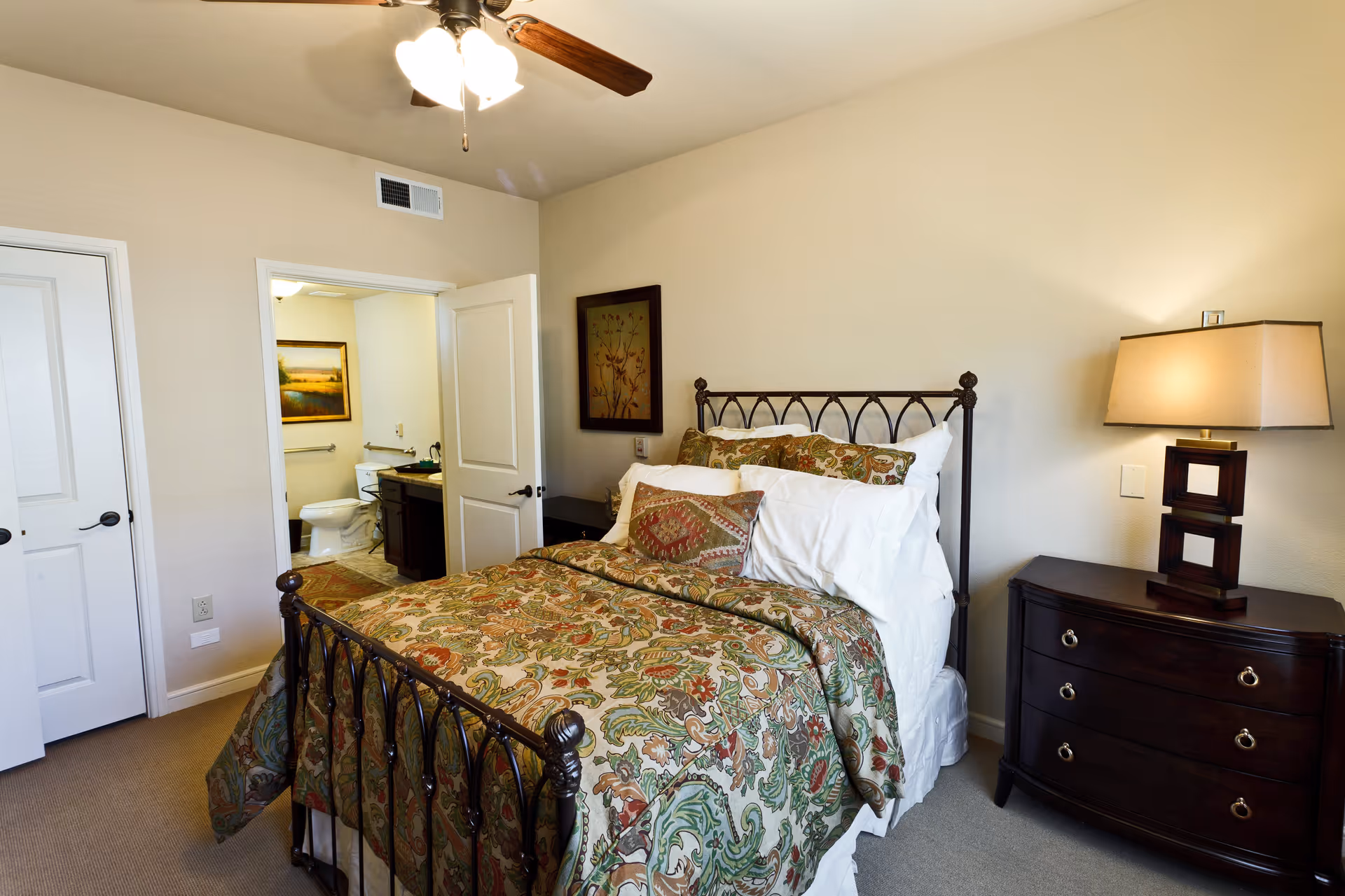 A bedroom with a decorative metal bed frame, a bed with patterned bedding and multiple pillows, a dark wooden nightstand with a lamp, and an open door leading to a bathroom with a toilet and sink visible. The walls are light-colored, and there is a ceiling fan with lights above.